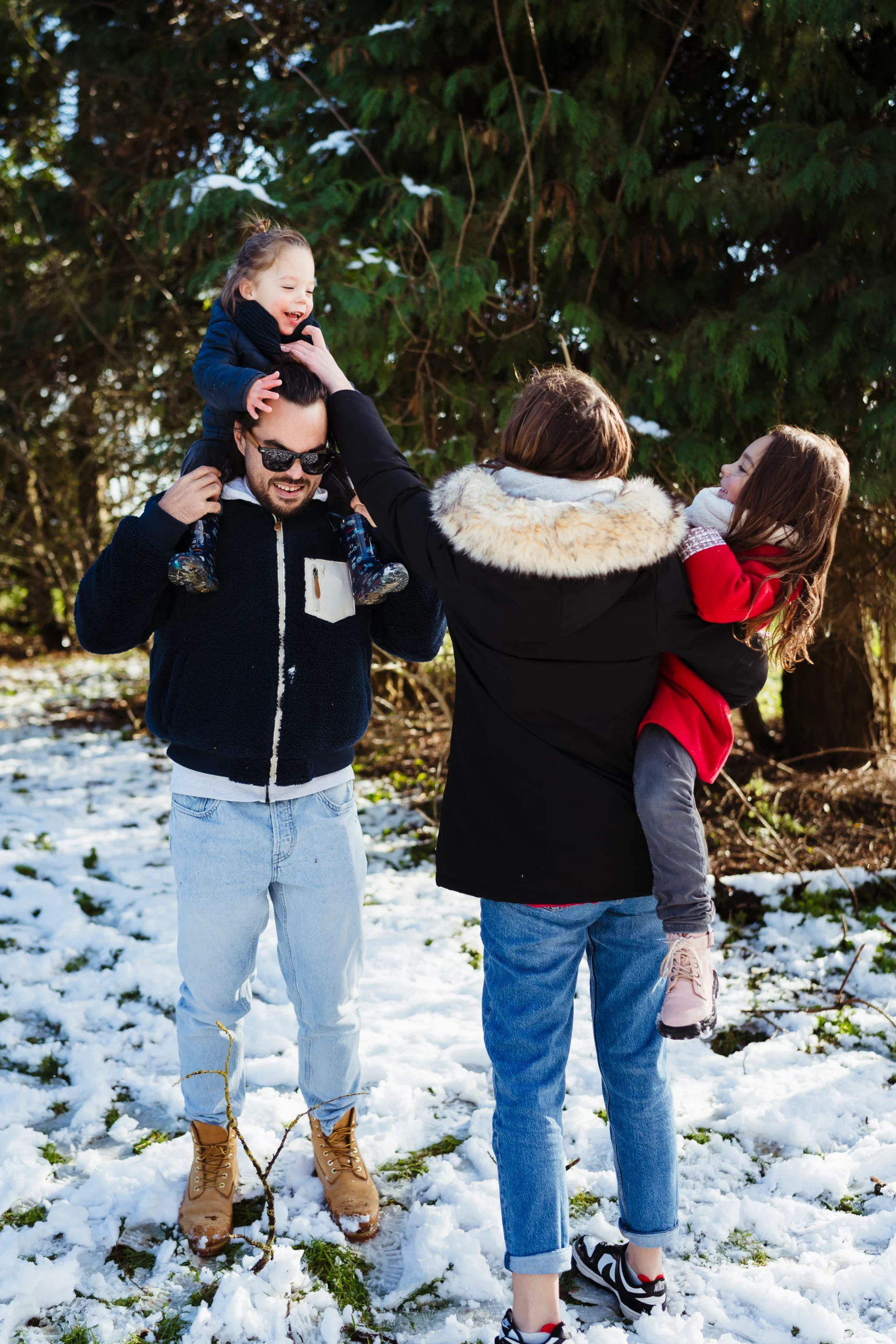 Portrait de famille d'un papa qui porte son fils sur les épaules et d'une maman qui serre sa fille dans les bras pour une séance photo de famille à fontainebleau en hiver